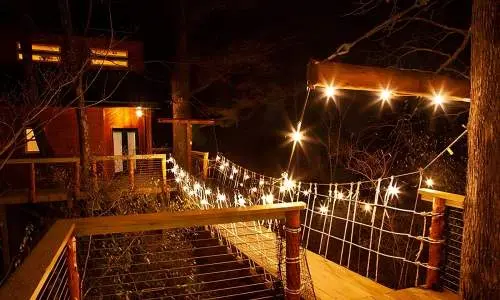 Un puente de madera hacia una cabaña en un árbol iluminada con luces de cuerda