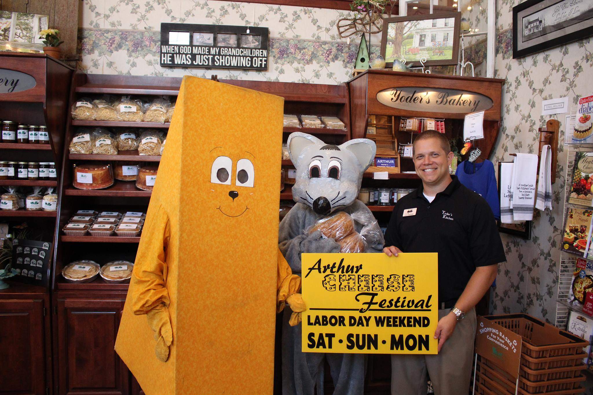 Mascotas disfrazadas de queso y ratón junto a un hombre que sostiene un cartel del Arthur Cheese Festival.