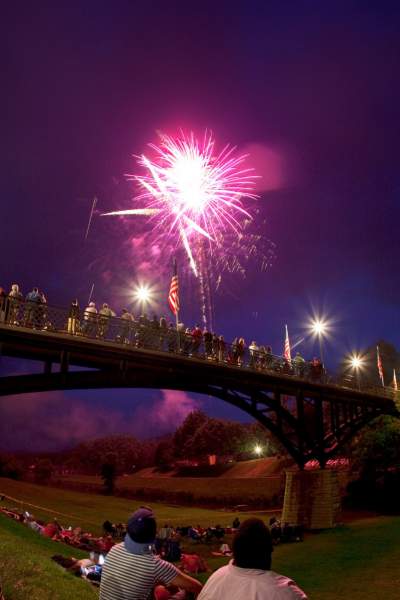 Gente sentada en un parque junto a un puente, viendo fuegos artificiales en el cielo