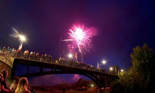 Gente sentada en un parque junto a un puente, viendo fuegos artificiales en el cielo