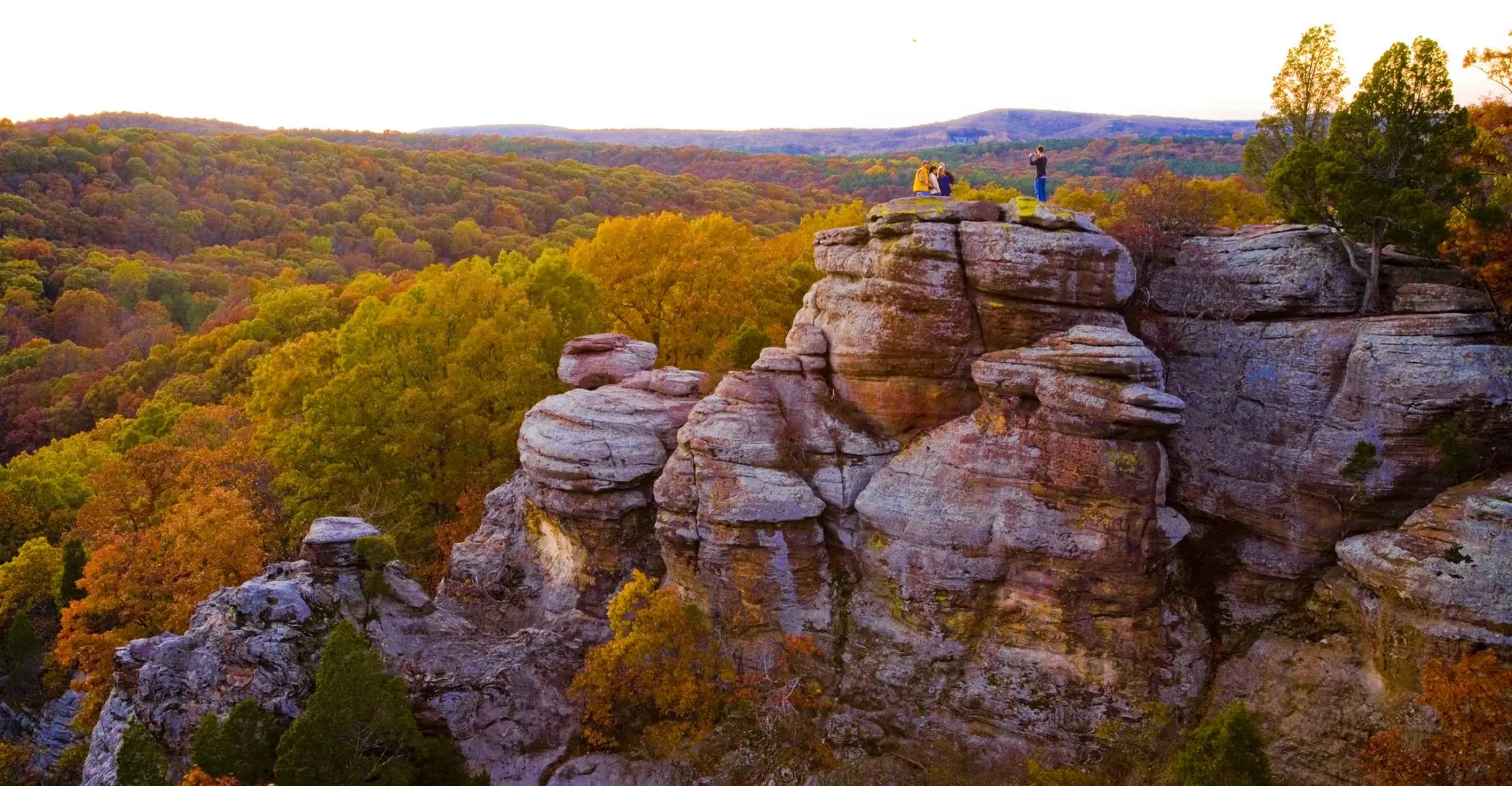 Un grupo distante de cuatro personas posando para una foto en lo alto de acantilados rocosos, con vistas a bosques de árboles verdes y dorados.