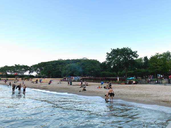 Pequeños grupos de personas en una tranquila playa junto a un lago al atardecer, con árboles bajos y verdes a sus espaldas.