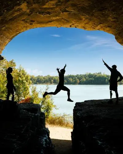 Una persona salta sobre una roca con amigos, cielo azul y agua de fondo