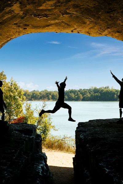 Una persona salta sobre una roca con amigos, cielo azul y agua de fondo