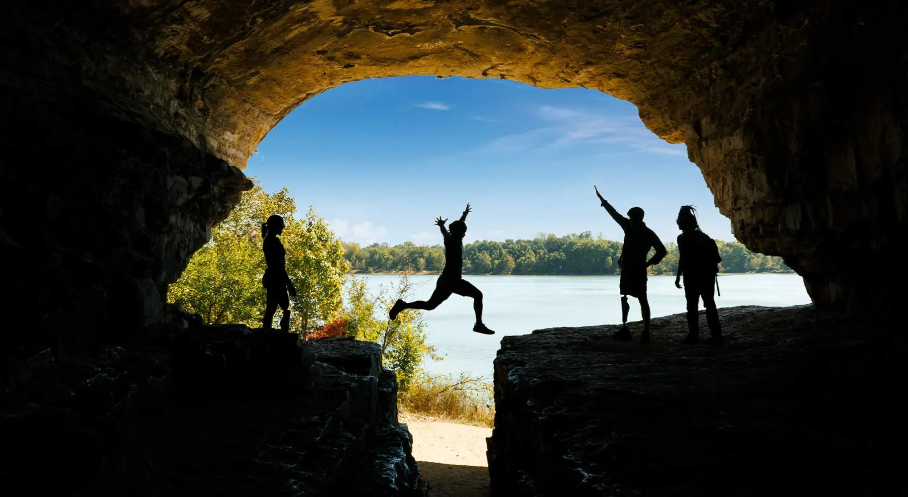 Una persona salta sobre una roca con amigos, cielo azul y agua de fondo
