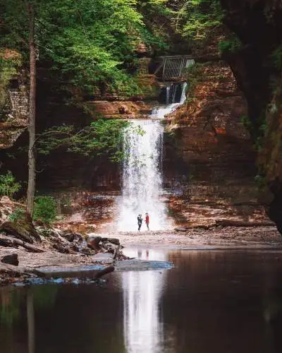 Dos personas de pie frente a una cascada, junto a un sereno estanque.