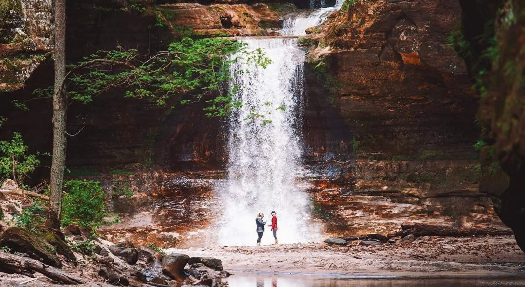 Dos personas de pie frente a una cascada, junto a un sereno estanque.