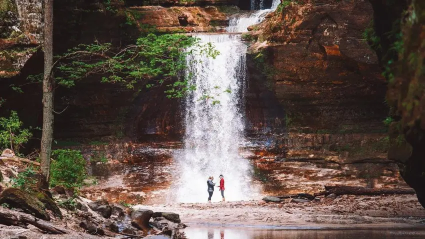 Dos personas de pie frente a una cascada, junto a un sereno estanque.