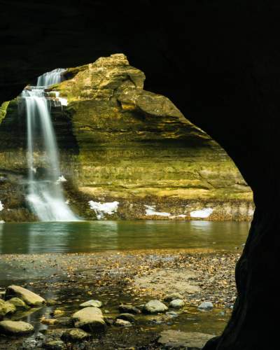 Mirando hacia el exterior desde una cueva, una cascada cae en un estanque, junto a paredes de roca revestidas de nieve