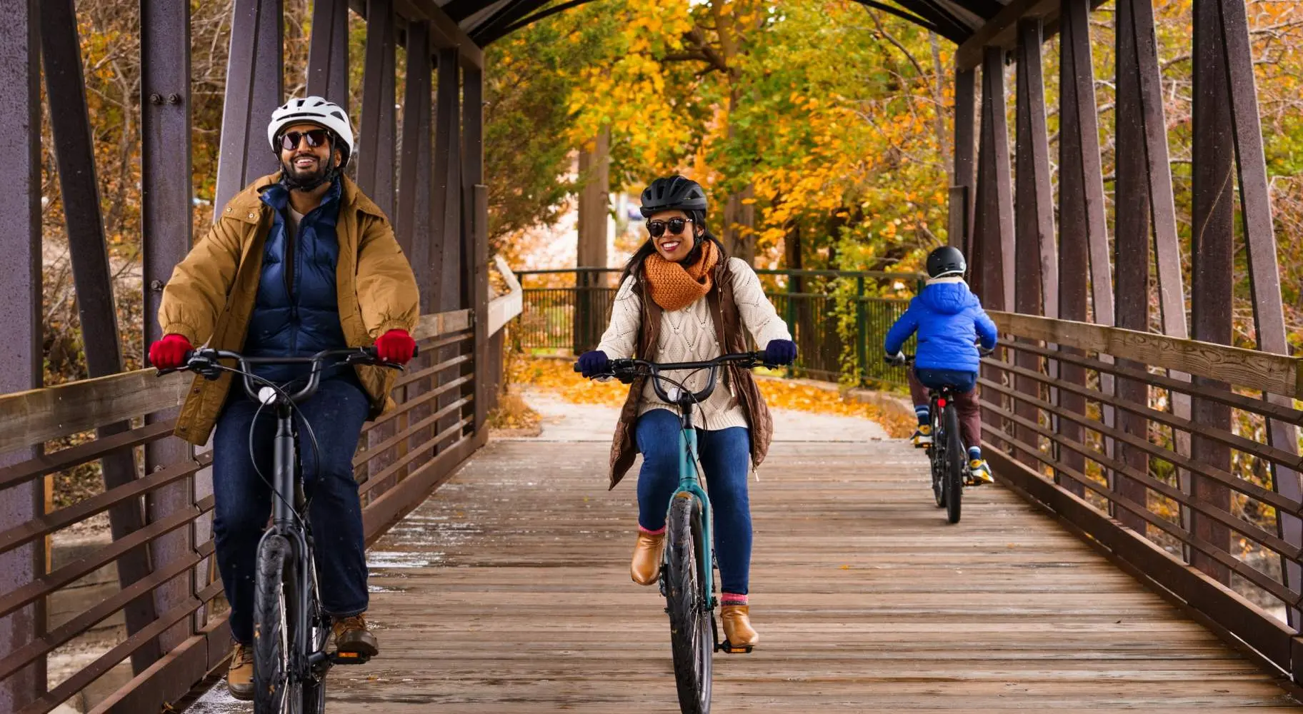 Una pareja pasea en bicicleta por un viejo puente cubierto de madera, con el follaje otoñal a sus espaldas