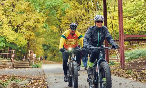 Dos ciclistas recorren un sendero arbolado bajo un viejo puente de madera
