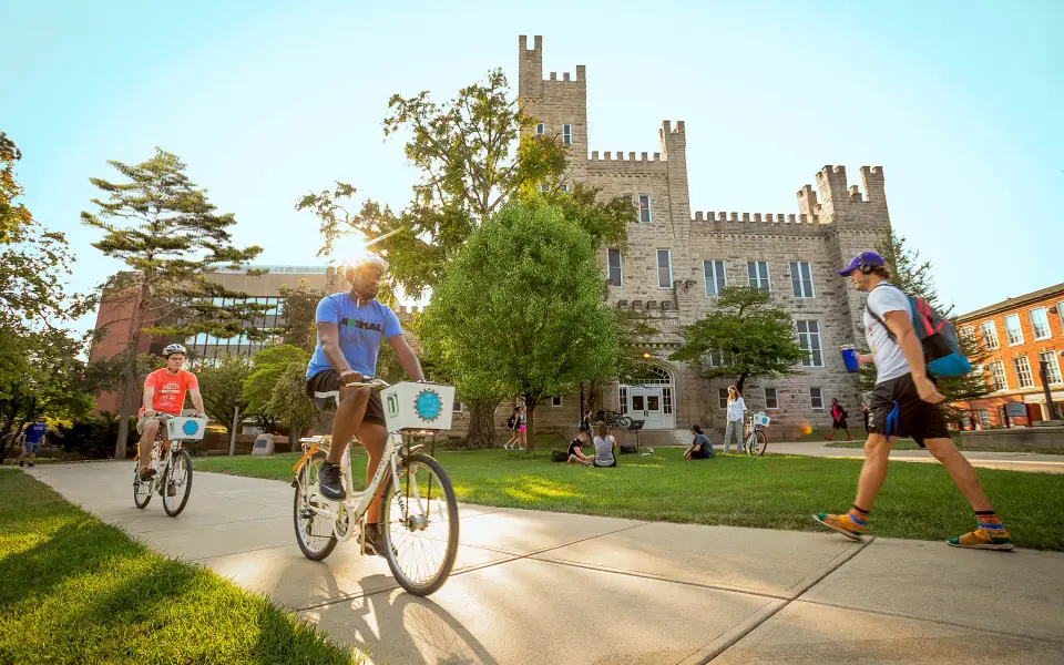 Dos personas recorren en bicicleta un campus universitario en un día soleado