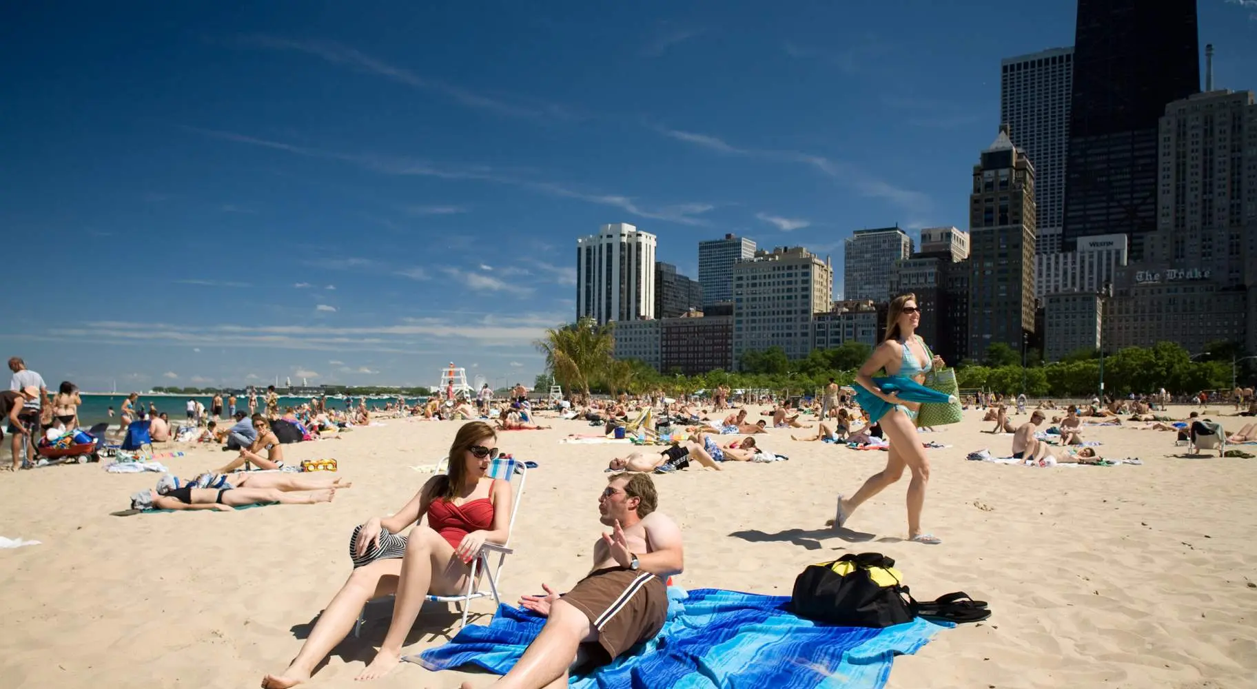 Dos personas tumbadas en una playa de arena, con multitudes al fondo, junto al horizonte de Chicago