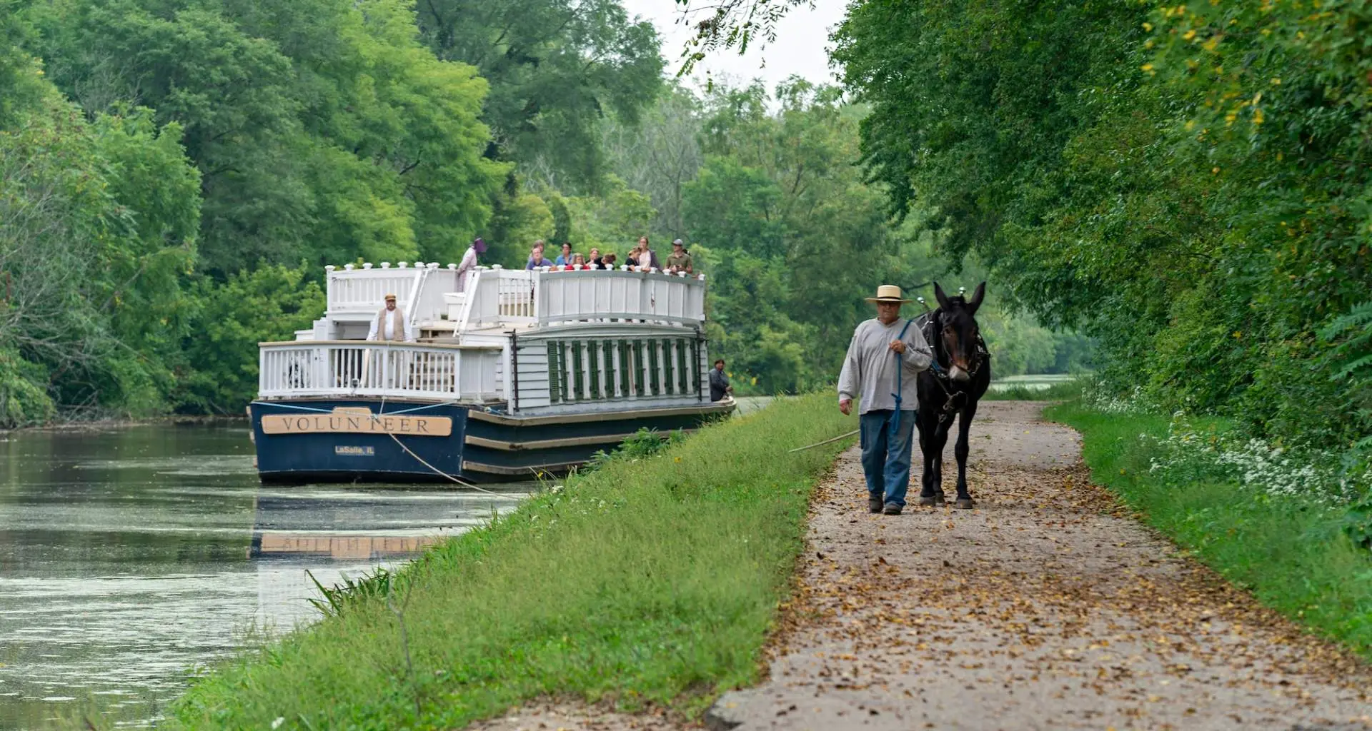 Un barco bajando por el río y un hombre paseando una mula por un camino