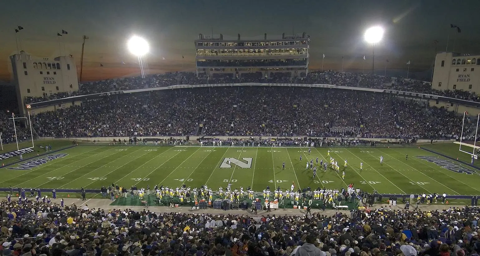 Un gran estadio de fútbol universitario por la noche antes de un partido, con las gradas llenas de espectadores