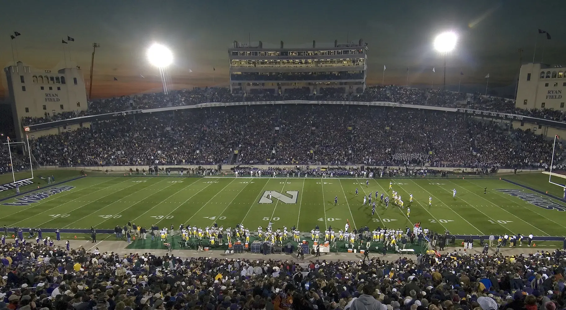Un gran estadio de fútbol universitario por la noche antes de un partido, con las gradas llenas de espectadores