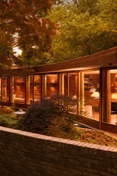 Una moderna casa de cristal con formas curvas, iluminada al atardecer, rodeada de árboles y un muro bajo de ladrillo.