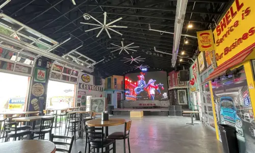 Interior de un bar espacioso y local de música en directo, con mesas, carteles y decoración de coches clásicos, y una gran pantalla sobre el escenario.